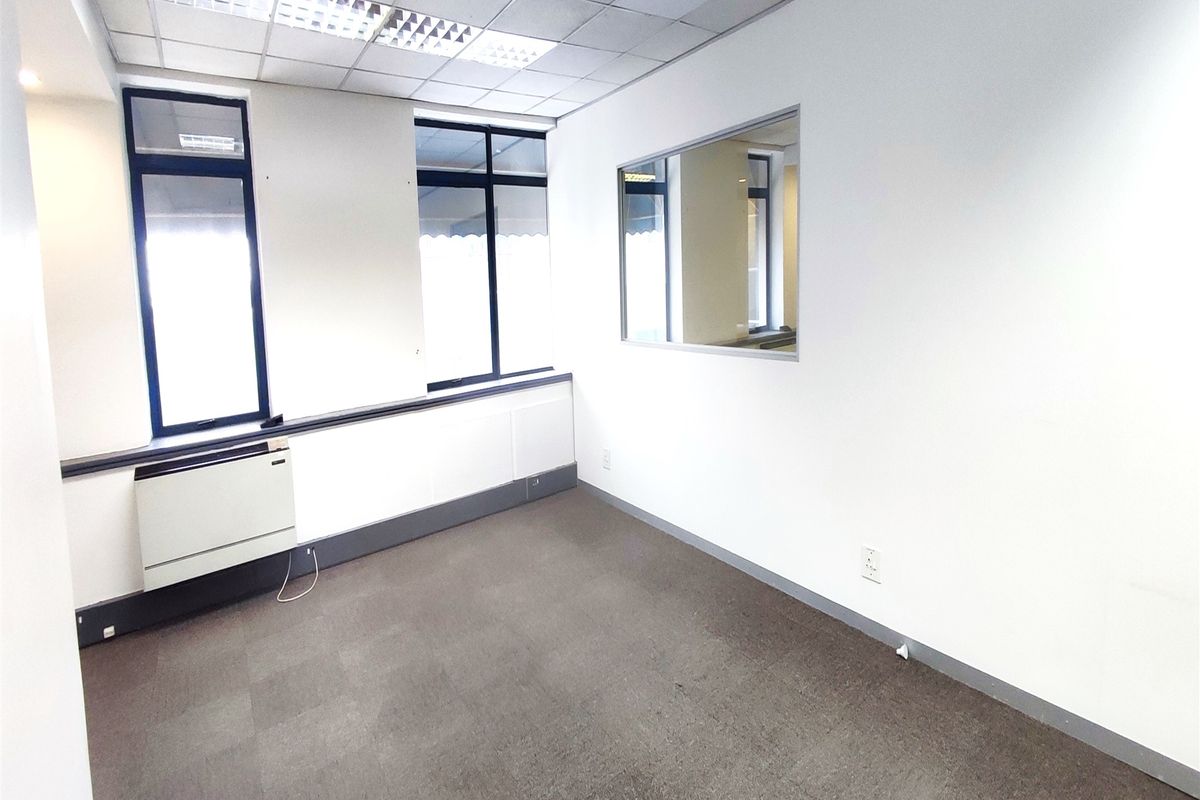 A clean, empty office space featuring carpet flooring, a suspended ceiling with recessed lighting, an internal window, and a wall-mounted HVAC unit.
