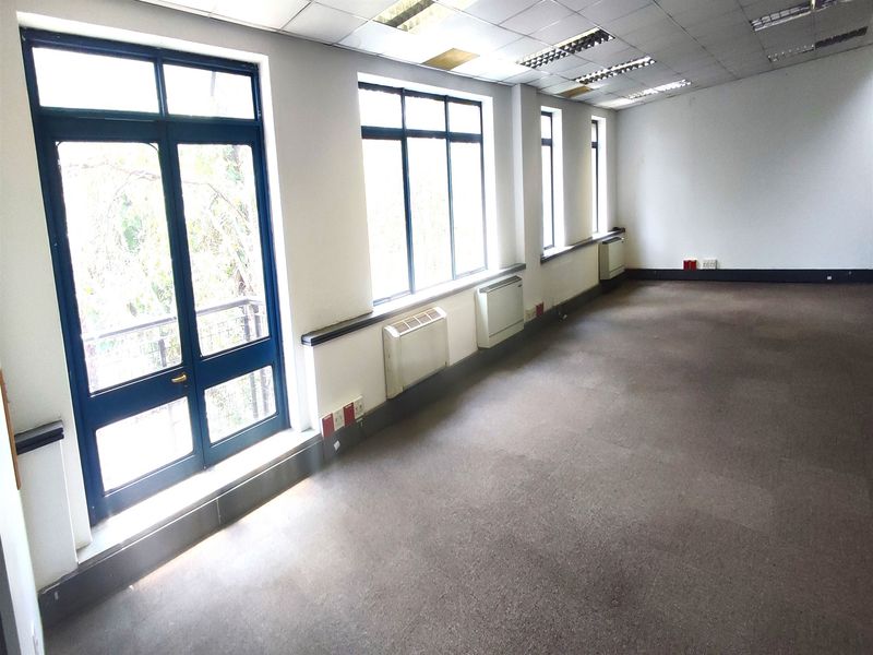 Spacious, empty office area featuring large windows, wall-mounted HVAC units, carpeted flooring, and a suspended ceiling with recessed lighting.