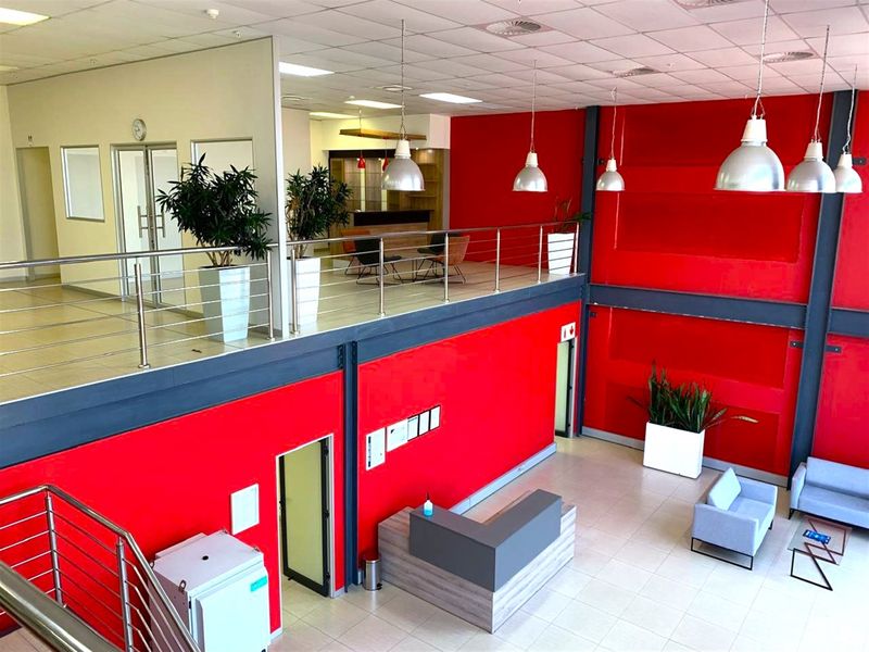 Spacious industrial open-plan office area featuring a mezzanine level, modern reception desk, high ceilings with pendant lighting, and vibrant red accent walls.