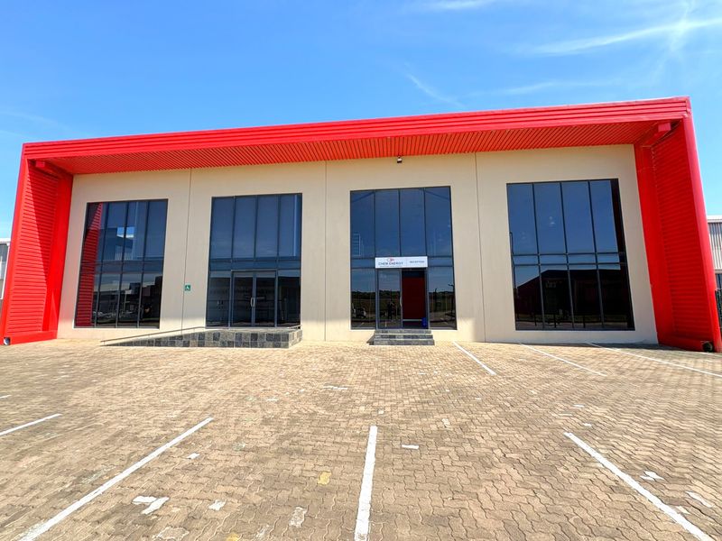 Modern industrial building facade featuring a prominent red metal roof, large glass windows, and a paved parking area with an accessible entrance ramp.