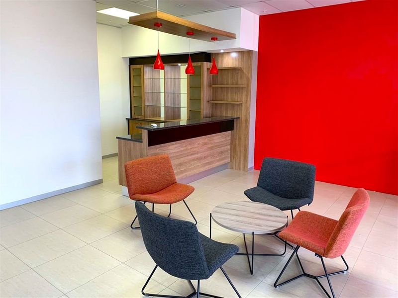 A modern office break area featuring a built-in bar counter with timber cabinetry, pendant lighting, and a vibrant red accent wall.
