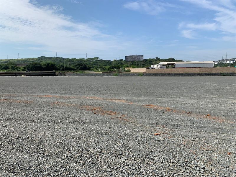 A wide, level commercial vacant land plot featuring a gravel surface, retaining wall, and distant views of surrounding landscape and infrastructure.