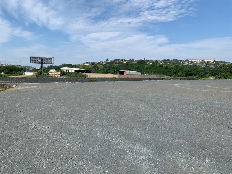 A wide-angle view of a vacant commercial land plot featuring a gravel surface, retaining wall, and an existing billboard structure.
