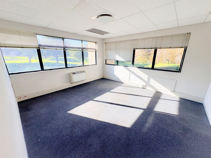 A bright, empty office space featuring large windows with venetian blinds, carpeted flooring, a suspended ceiling, and a wall-mounted air conditioning unit.