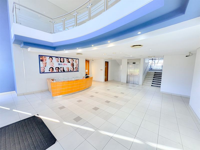Spacious office building entrance lobby featuring a reception desk, tiled flooring, stainless steel elevator, and a mezzanine level with glass balustrade.