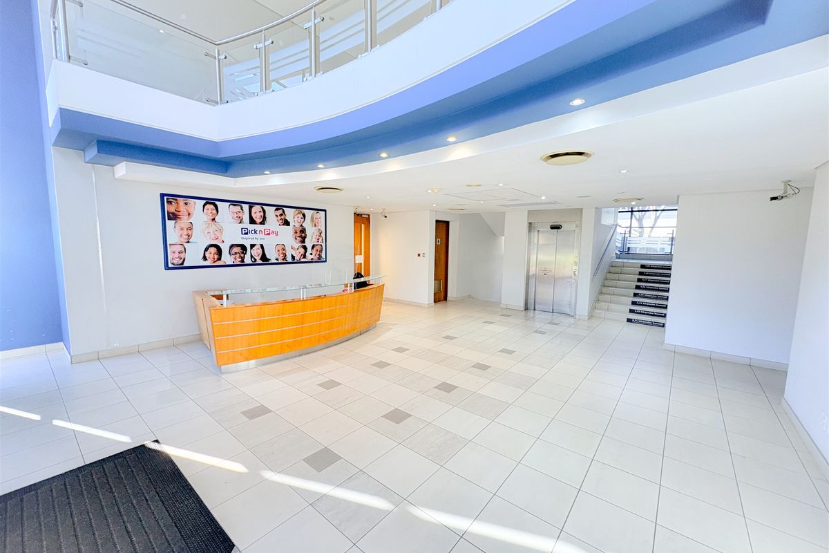 Spacious office building entrance lobby featuring a reception desk, tiled flooring, stainless steel elevator, and a mezzanine level with glass balustrade.