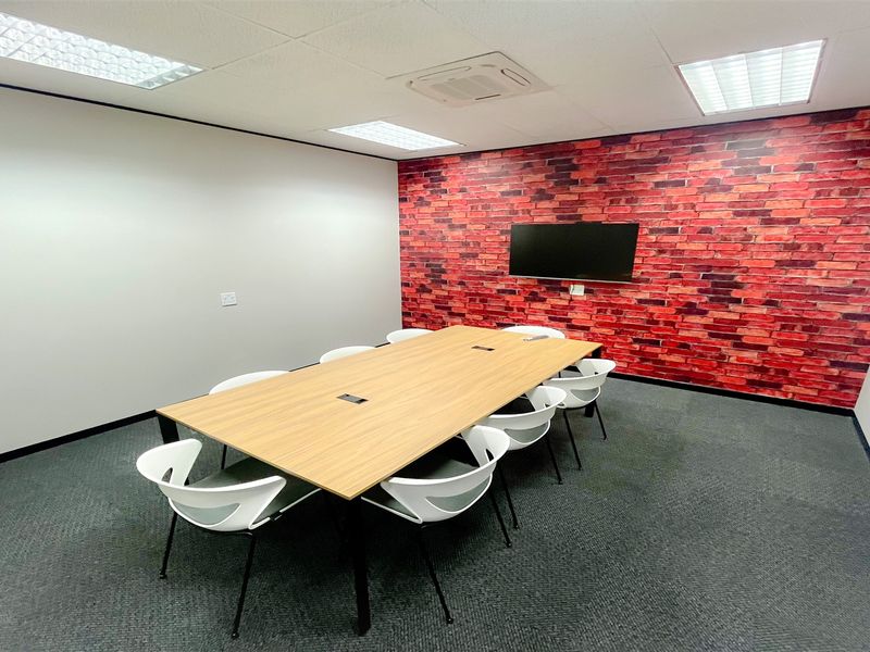 A professional office meeting room featuring a modern wood-finish conference table, carpeted flooring, recessed lighting, and a decorative brick feature wall.