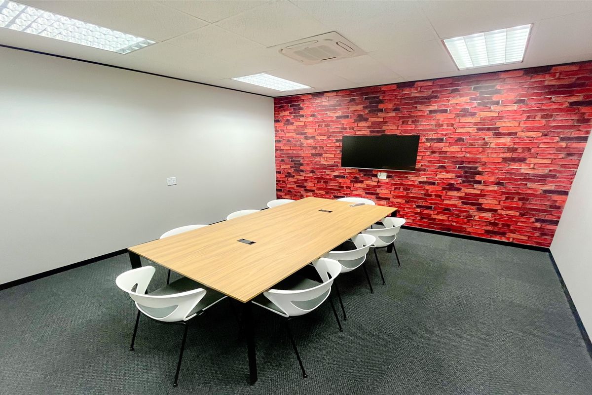 A professional office meeting room featuring a modern wood-finish conference table, carpeted flooring, recessed lighting, and a decorative brick feature wall.