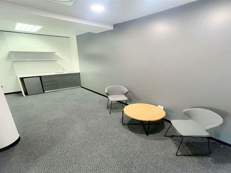A modern office space featuring a compact kitchenette with built-in cabinetry, sink, and recessed lighting, providing a functional break area.