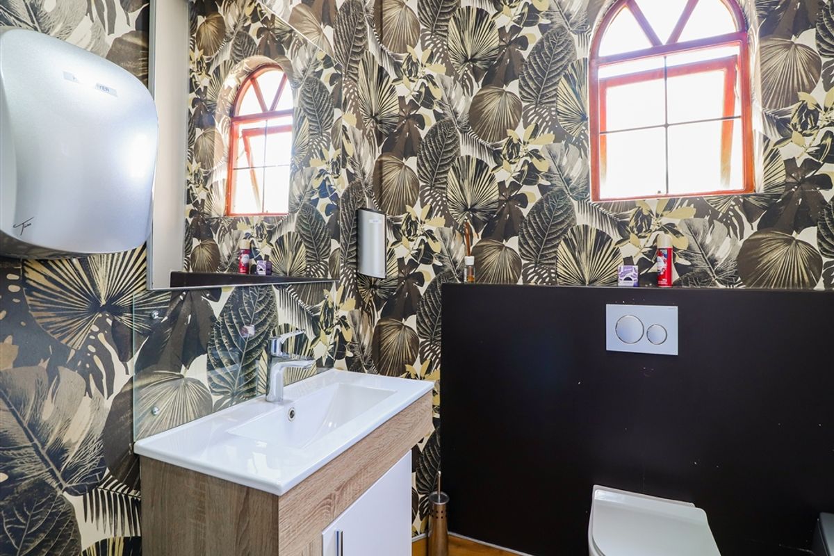 Modern commercial bathroom featuring a vanity with basin, wall-hung toilet with concealed cistern, and decorative botanical wallpaper under arched windows.