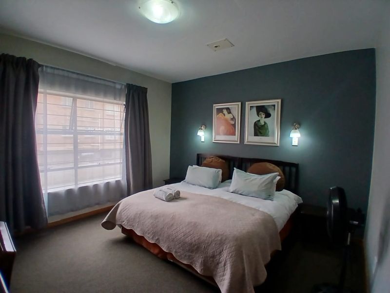 A bedroom featuring a dark accent wall, wall-mounted bedside lighting, and a large window with curtains.