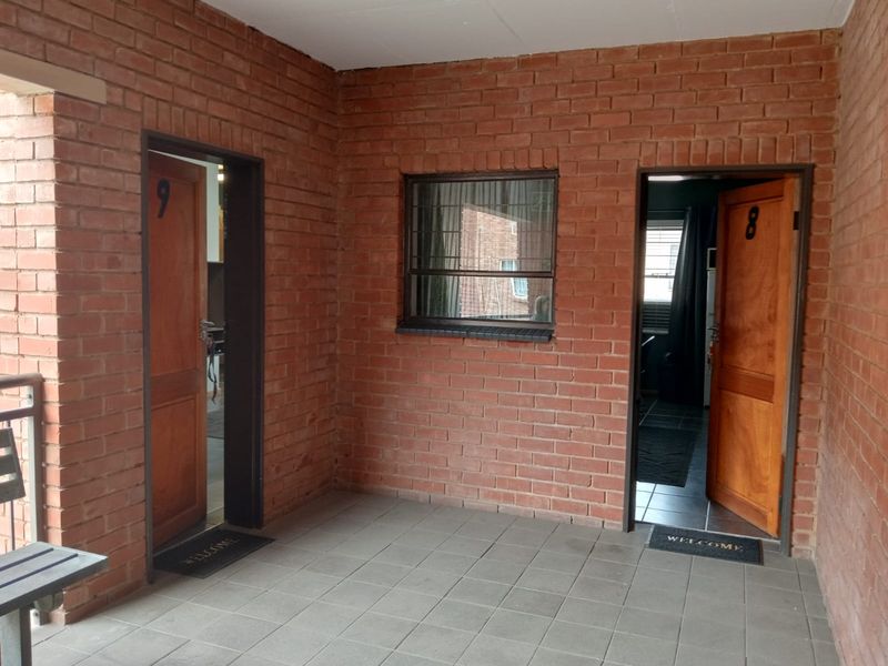 A covered exterior walkway featuring exposed brick walls, tiled flooring, and two wooden entrance doors leading into commercial units.