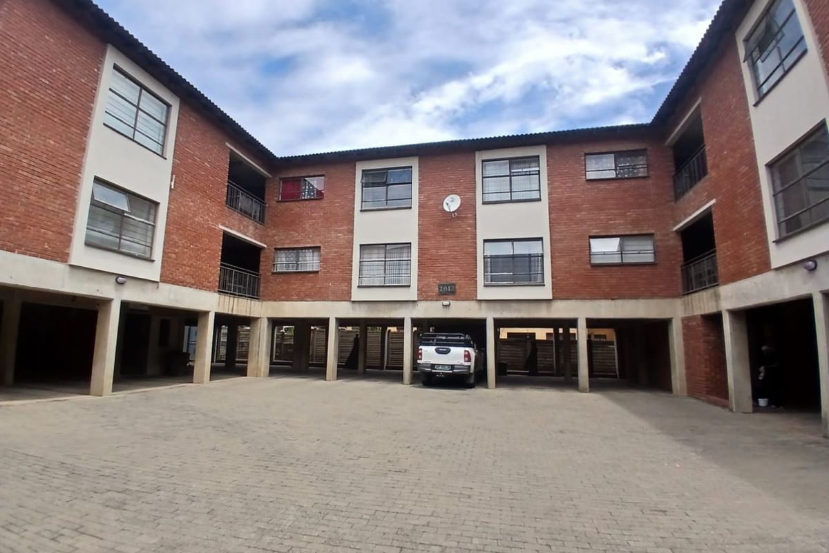 Exterior view of a multi-story commercial or residential complex featuring exposed brick facade, covered ground-level parking area, and paved courtyard.