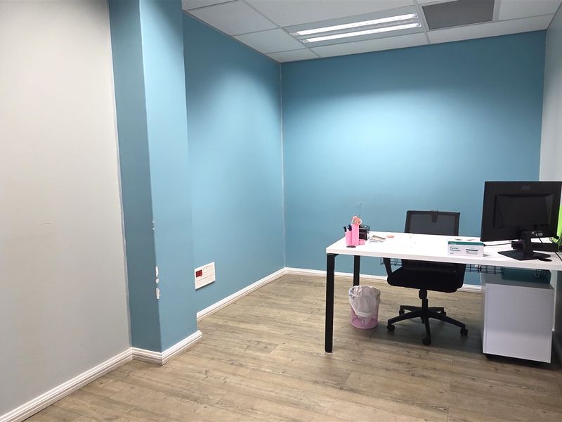 A compact office space featuring blue accent walls, wood-look laminate flooring, and a suspended ceiling with integrated lighting.