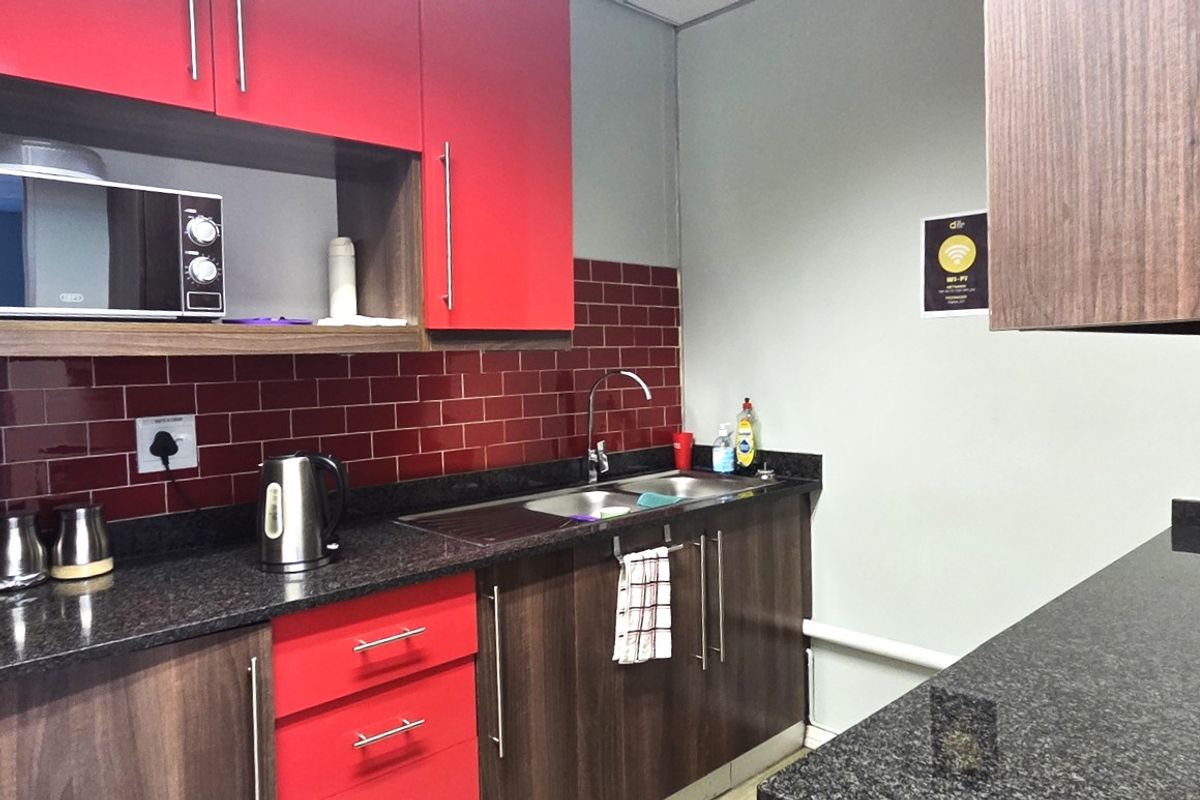 A compact office kitchenette featuring modern wood-grain and red cabinetry, granite countertops, a stainless steel sink, and a tiled backsplash.