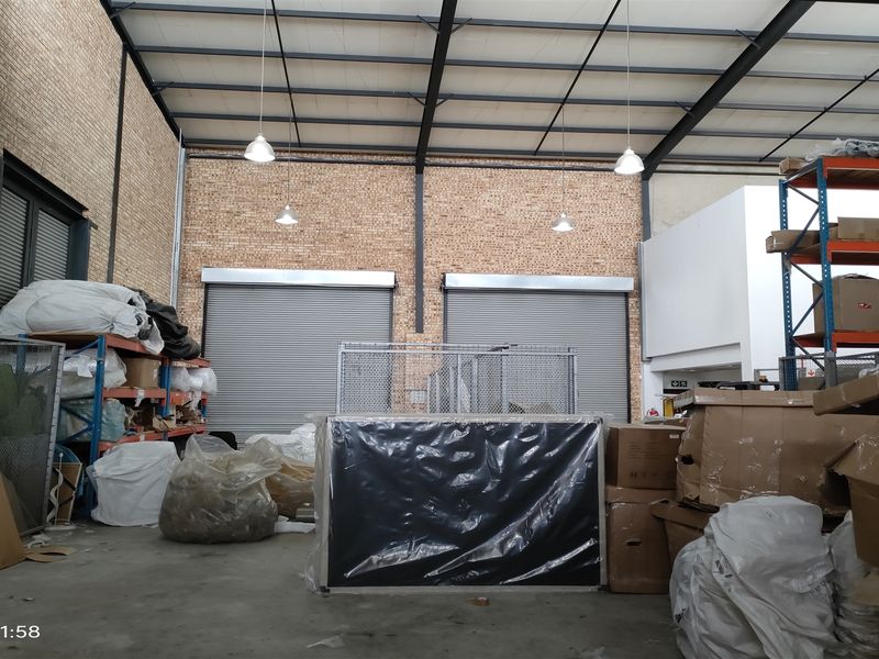 Industrial warehouse space featuring exposed brick walls, high steel truss ceilings, concrete flooring, and dual roller shutter doors for efficient access.