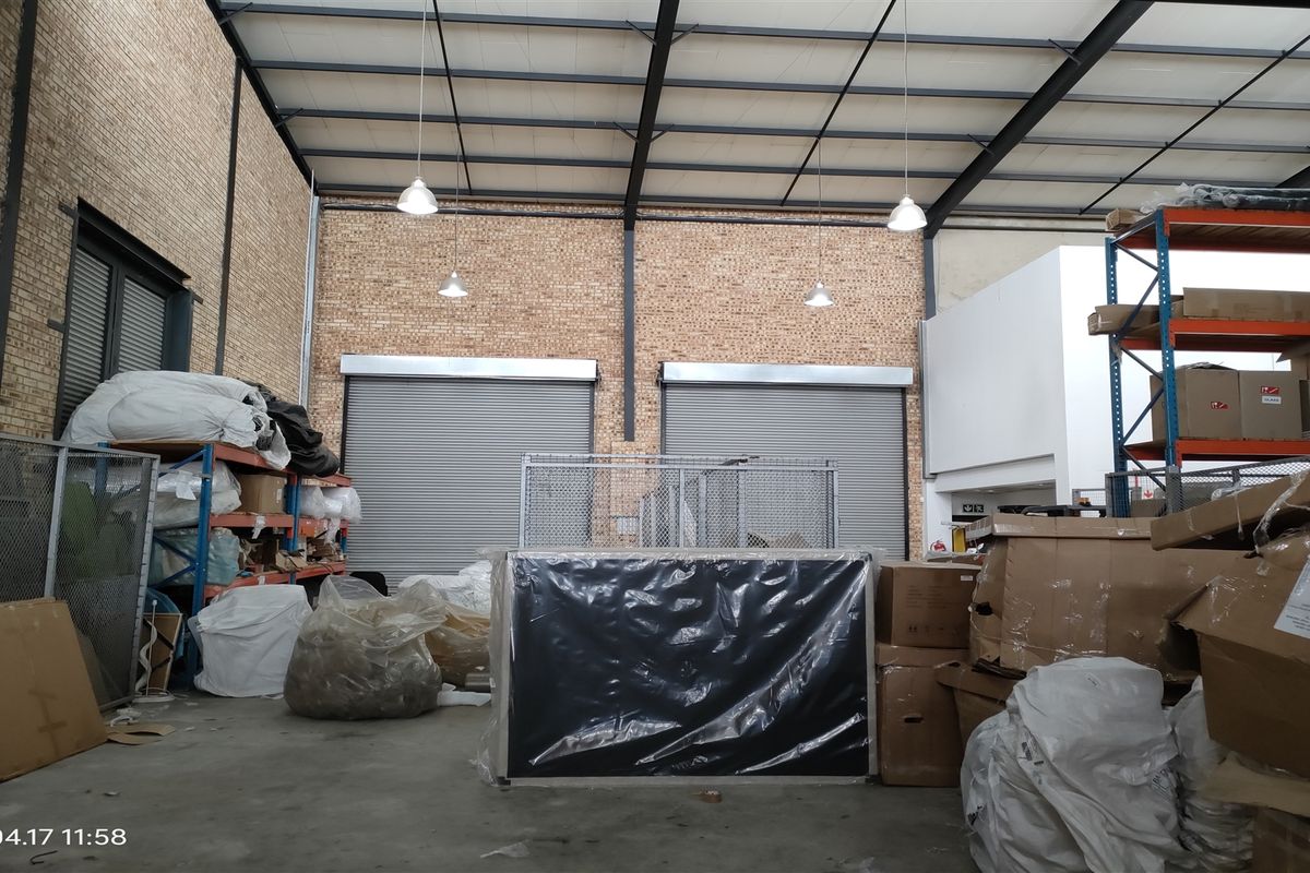 Industrial warehouse space featuring exposed brick walls, high steel truss ceilings, concrete flooring, and dual roller shutter doors for efficient access.
