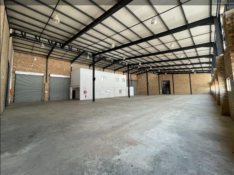 Spacious industrial warehouse featuring high ceilings with exposed steel trusses, concrete flooring, multiple roller shutter doors, and exposed brick wall finishes.
