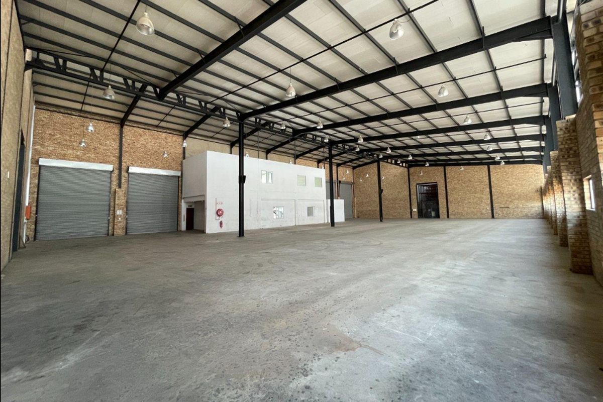 Spacious industrial warehouse featuring high ceilings with exposed steel trusses, concrete flooring, multiple roller shutter doors, and exposed brick wall finishes.
