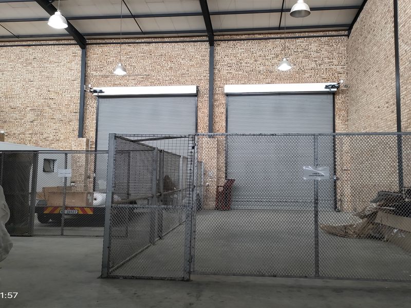 Industrial warehouse space featuring high ceilings with exposed steel trusses, face brick walls, concrete flooring, and dual roller shutter doors for loading access.