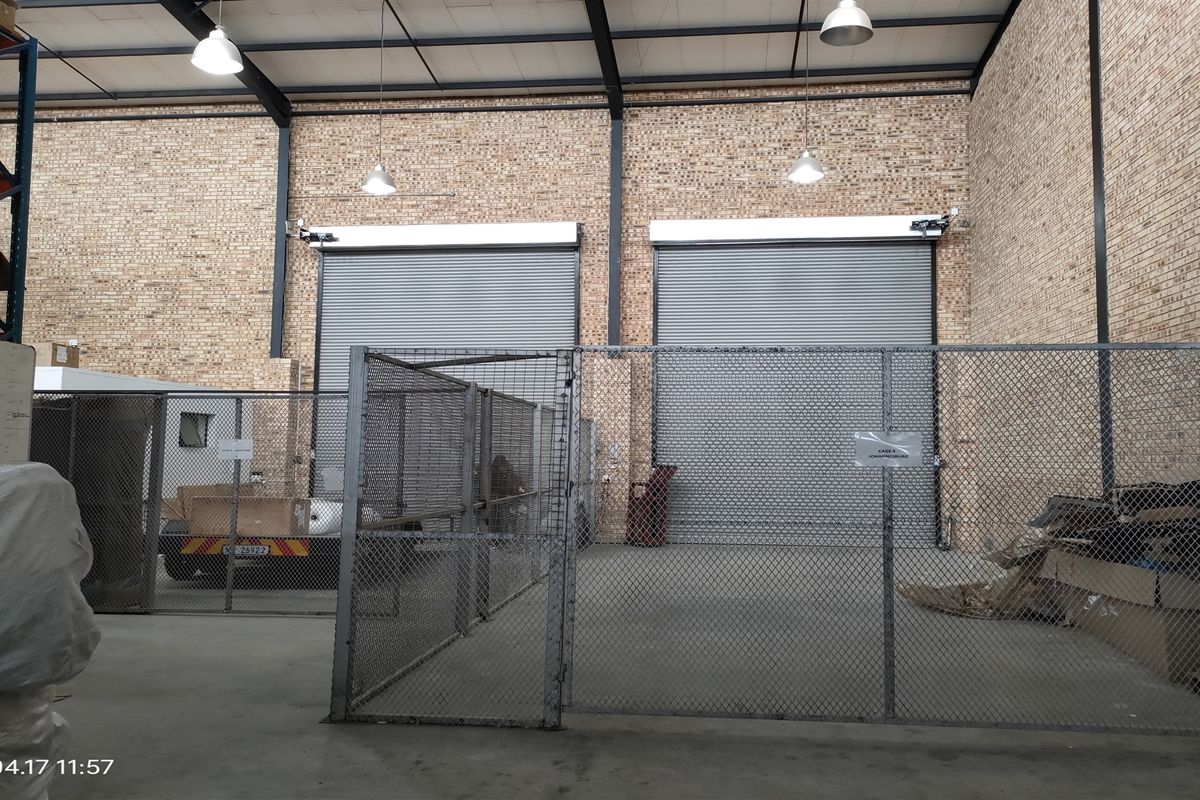 Industrial warehouse space featuring high ceilings with exposed steel trusses, face brick walls, concrete flooring, and dual roller shutter doors for loading access.