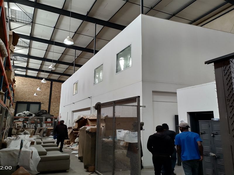 Industrial warehouse space featuring high ceilings with exposed steel trusses, a mezzanine level, metal security partitioning, and integrated industrial racking systems.