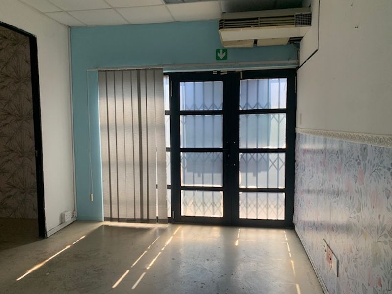 An empty office space featuring a glass door with security gate, vertical blinds, and a wall-mounted air conditioning unit.