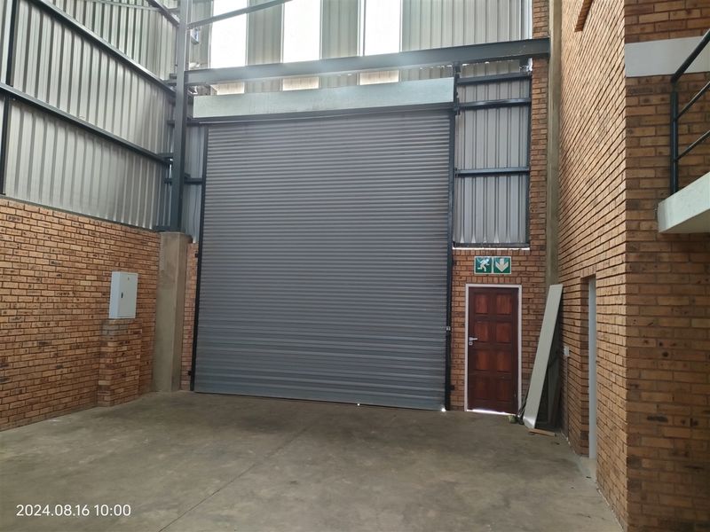 Industrial warehouse space featuring high ceilings, exposed brickwork, a large roller shutter door, and a separate pedestrian access door.