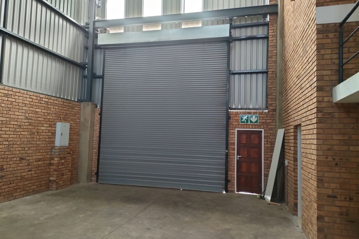 Industrial warehouse space featuring high ceilings, exposed brickwork, a large roller shutter door, and a separate pedestrian access door.