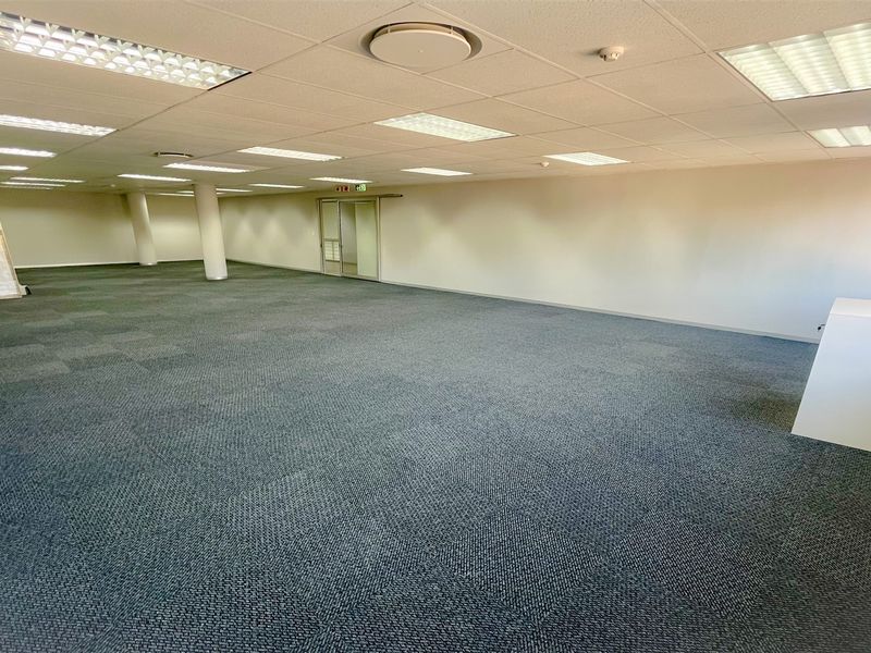 A spacious, open-plan office area featuring neutral walls, carpeted flooring, and a suspended ceiling with integrated recessed lighting.