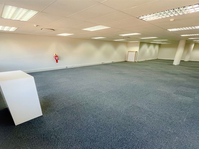 A spacious, open-plan office area featuring carpeted flooring, a suspended ceiling with recessed lighting, and a structural support column.