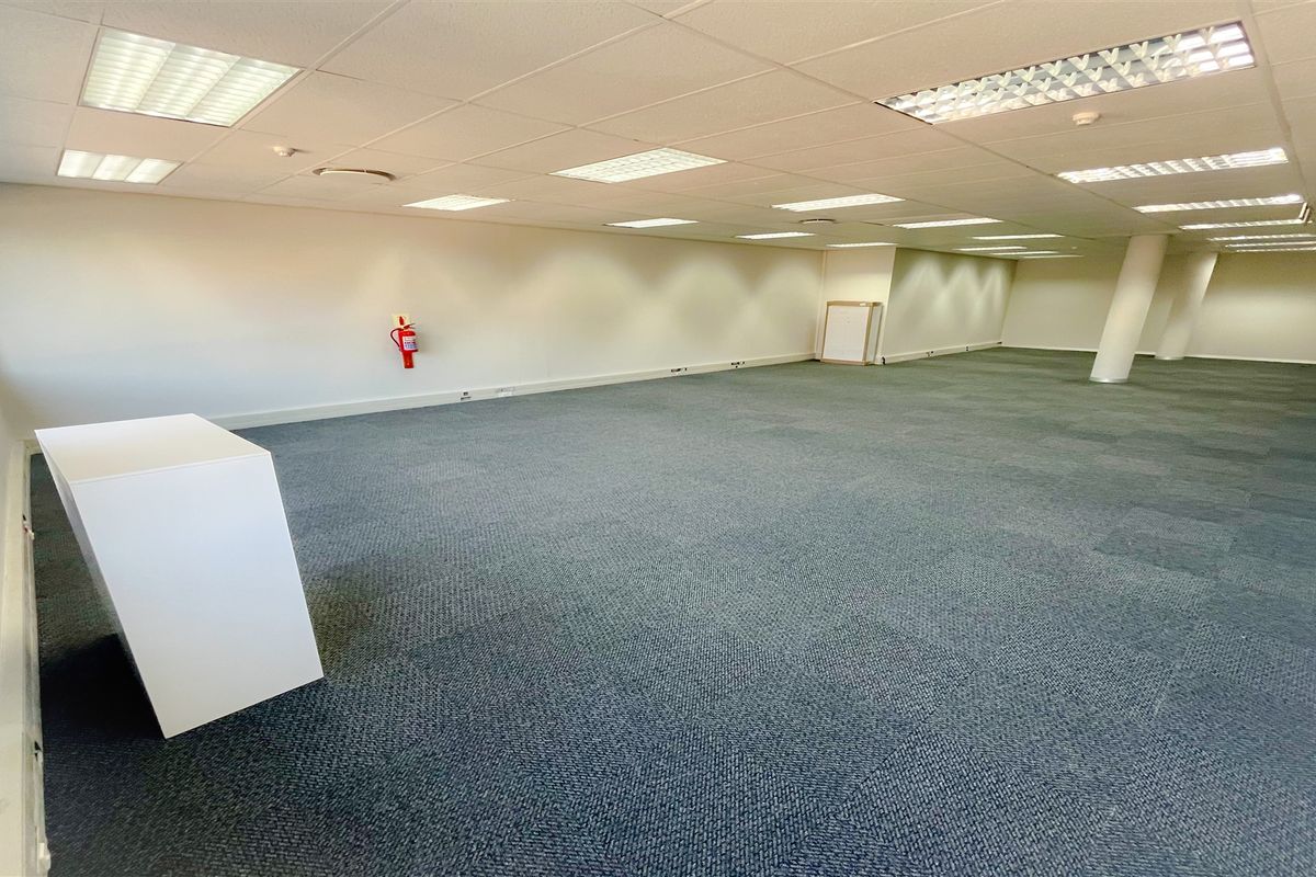 A spacious, open-plan office area featuring carpeted flooring, a suspended ceiling with recessed lighting, and a structural support column.