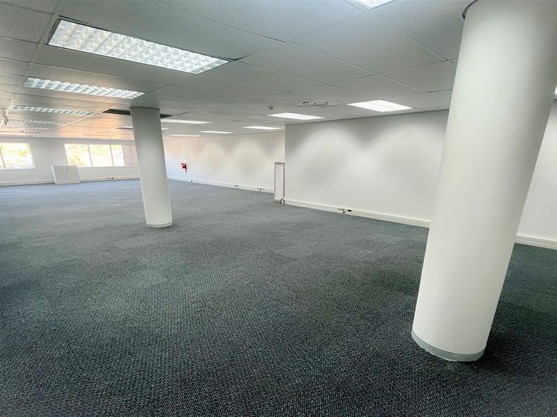 A spacious, open-plan office area featuring neutral carpet tiles, structural columns, and a suspended ceiling with recessed lighting.