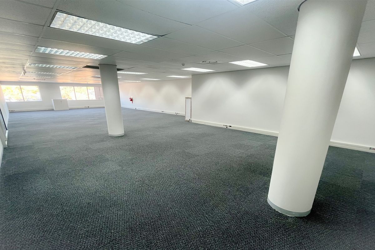 A spacious, open-plan office area featuring neutral carpet tiles, structural columns, and a suspended ceiling with recessed lighting.