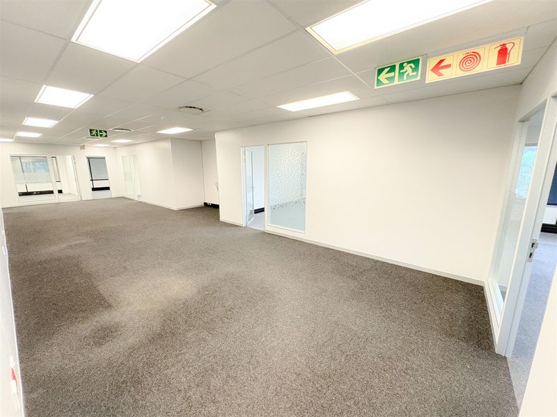 A spacious, vacant open-plan office area featuring neutral carpeting, suspended ceilings with recessed lighting, and glass-partitioned office suites.