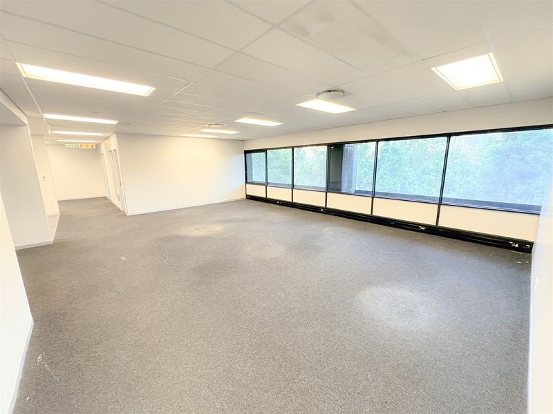 A spacious, empty open-plan office area featuring a suspended ceiling, recessed lighting, and a large window wall providing natural light.