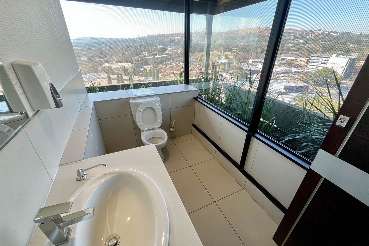 Modern office bathroom featuring a vanity basin, toilet, and expansive floor-to-ceiling windows offering panoramic views of the surrounding area.