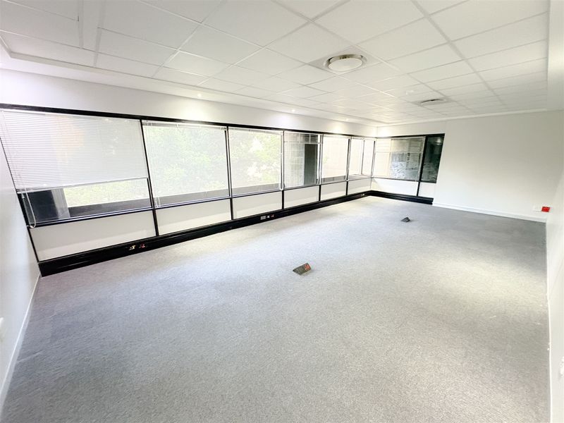 A bright, open-plan office space featuring a suspended ceiling, large windows with fitted blinds, and carpeted flooring with integrated floor power outlets.