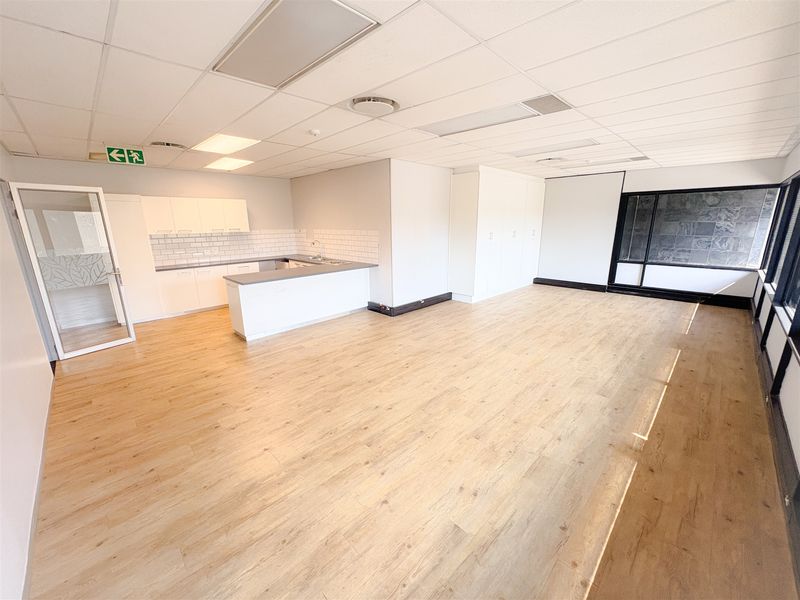 A bright, open-plan office space featuring light wood-look laminate flooring, a modern kitchenette with subway tile backsplash, and ample built-in storage cupboards.