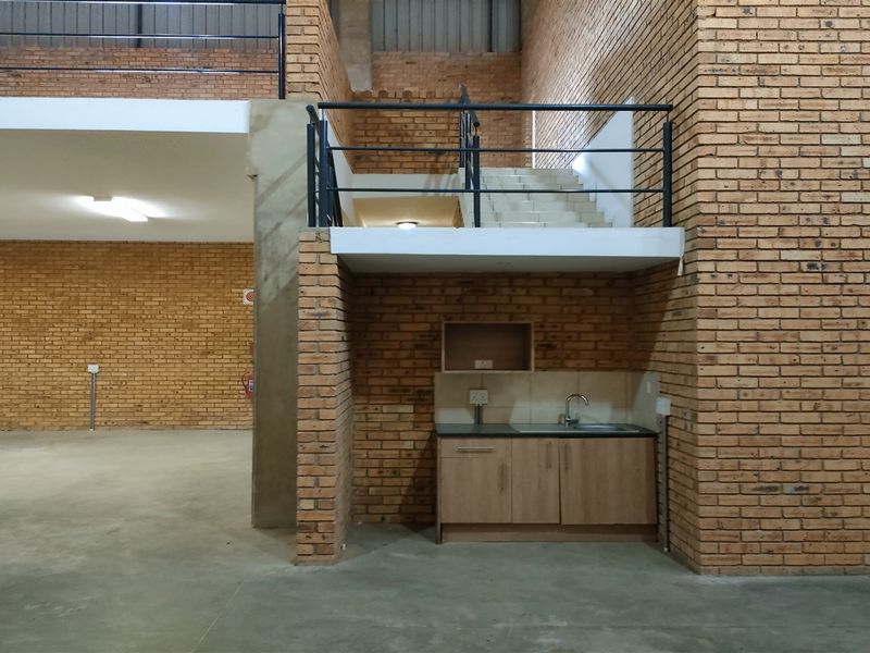 Spacious industrial open-plan area featuring exposed brick walls, a mezzanine level with metal staircase, and a functional kitchenette with sink.
