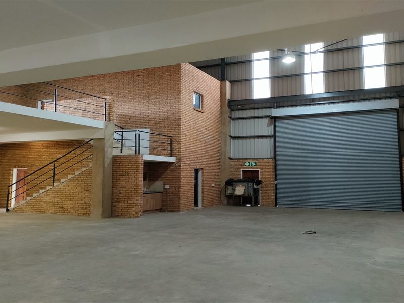 Spacious industrial warehouse unit featuring a large roller shutter door, mezzanine level, exposed brick walls, and high ceilings with integrated lighting.