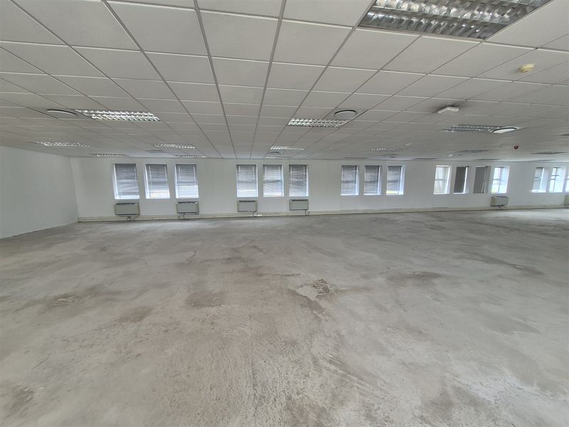 A large, open-plan office space featuring a suspended ceiling, recessed lighting, and wall-mounted air conditioning units, with unfinished concrete flooring.