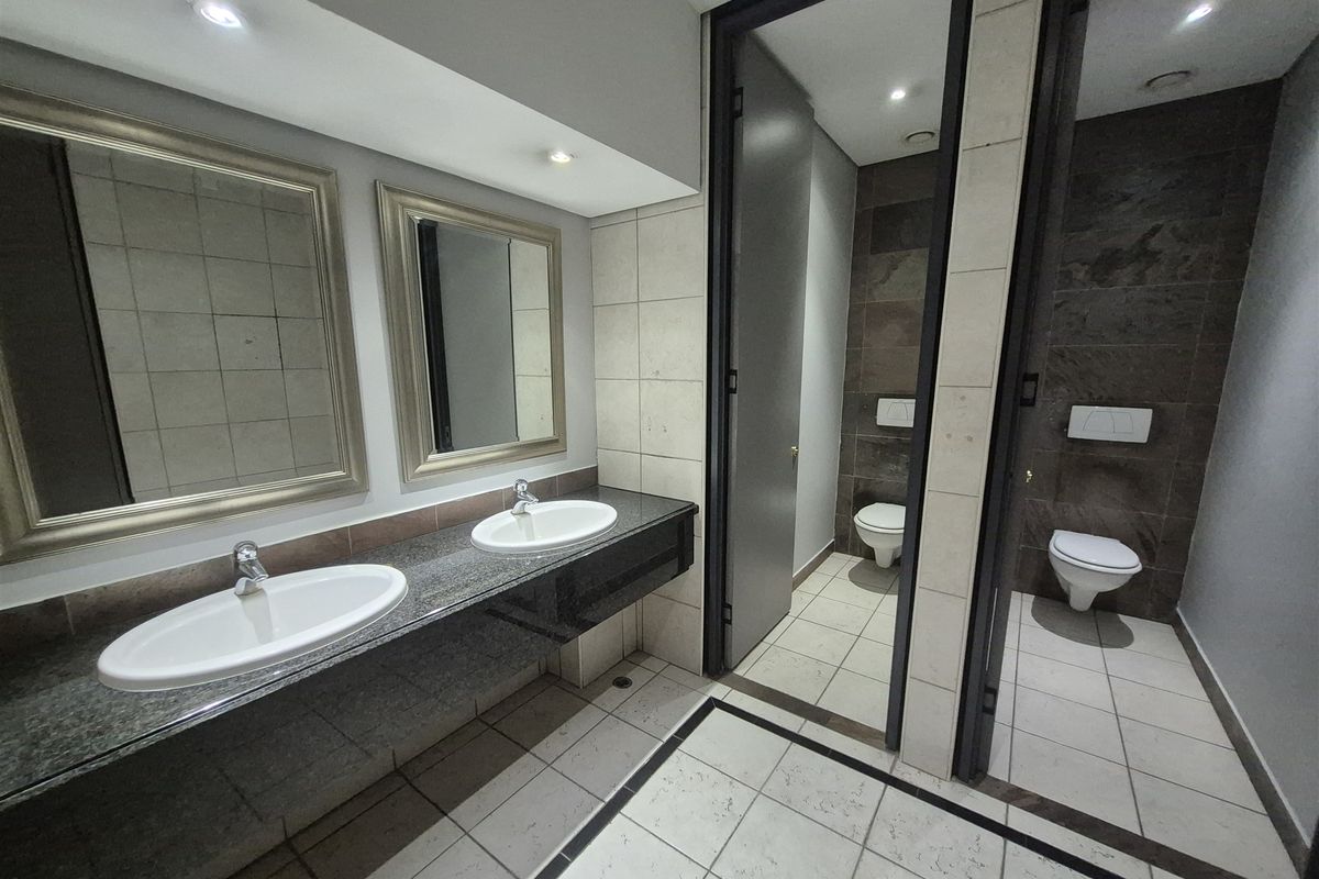 A modern, well-maintained office bathroom featuring a double vanity with granite countertops and two separate toilet cubicles with tiled finishes.