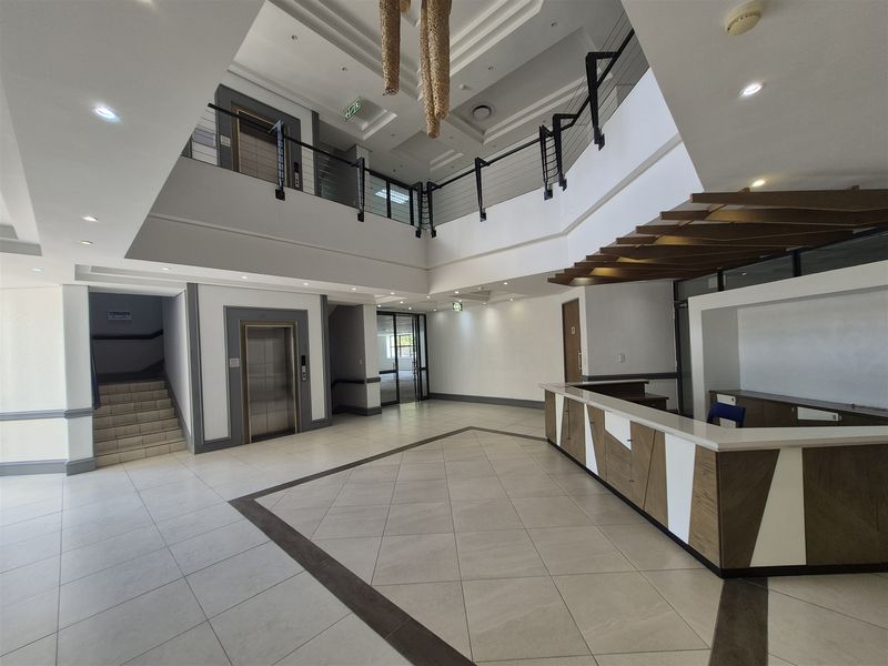 Spacious double-volume office building entrance lobby featuring a built-in reception desk, elevator access, and a mezzanine level with modern tiled flooring.