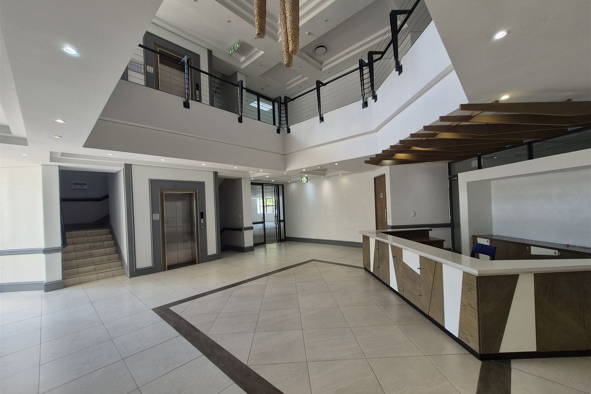 Spacious double-volume office building entrance lobby featuring a built-in reception desk, elevator access, and a mezzanine level with modern tiled flooring.