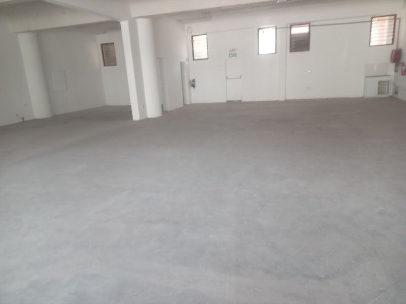 A spacious, open-plan retail unit featuring concrete flooring, structural columns, multiple windows, and a dedicated exit door.