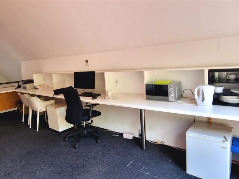 An office space featuring a long built-in desk with integrated shelving, situated under a sloped ceiling with carpeted flooring.