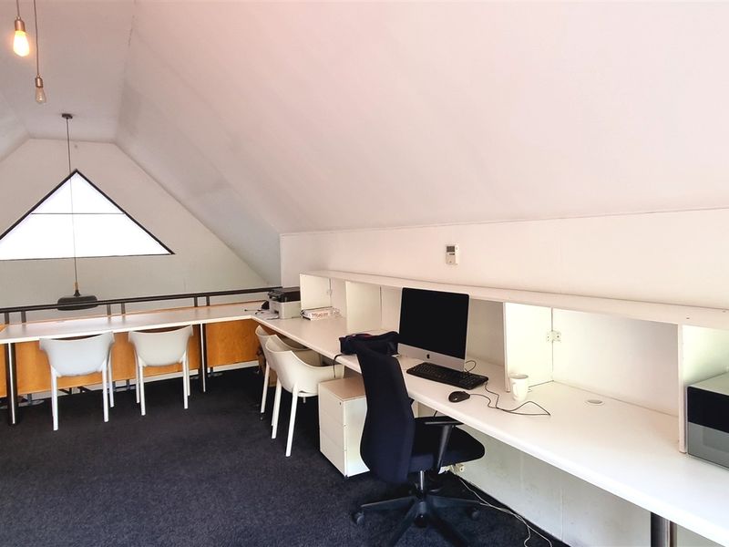 Spacious attic-style office featuring a long built-in desk, triangular window, and mezzanine layout with carpeted flooring.