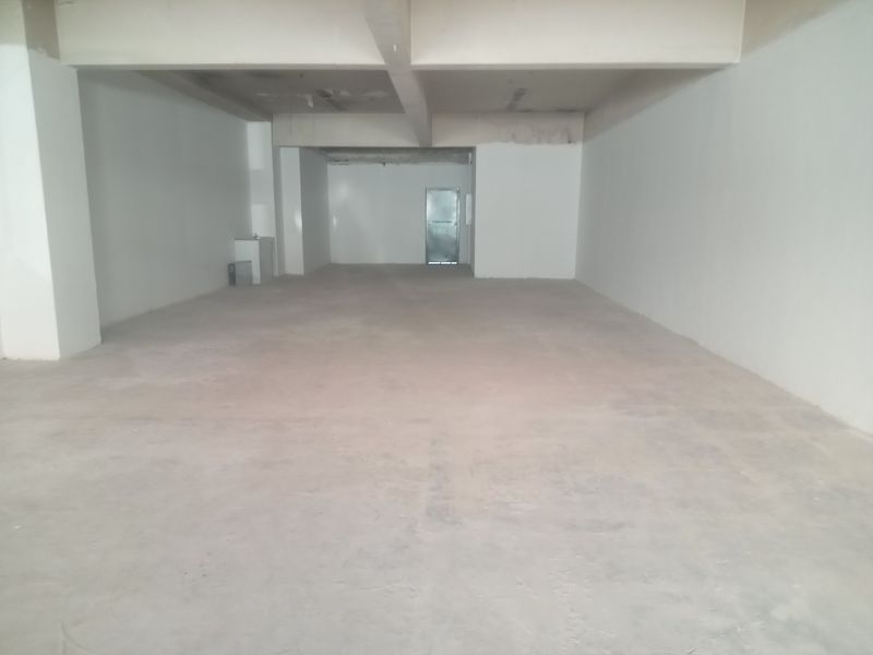 An open-plan retail space in shell condition, featuring concrete flooring, exposed structural beams, and a rear access door.