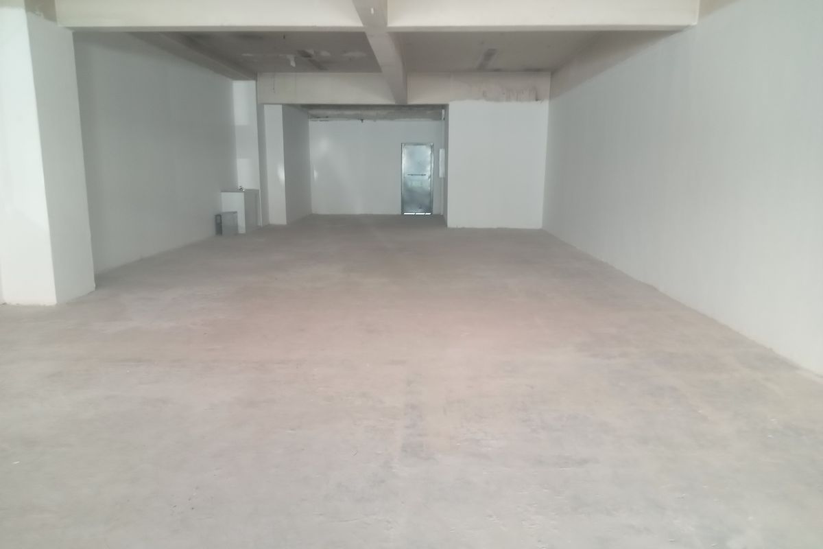 An open-plan retail space in shell condition, featuring concrete flooring, exposed structural beams, and a rear access door.